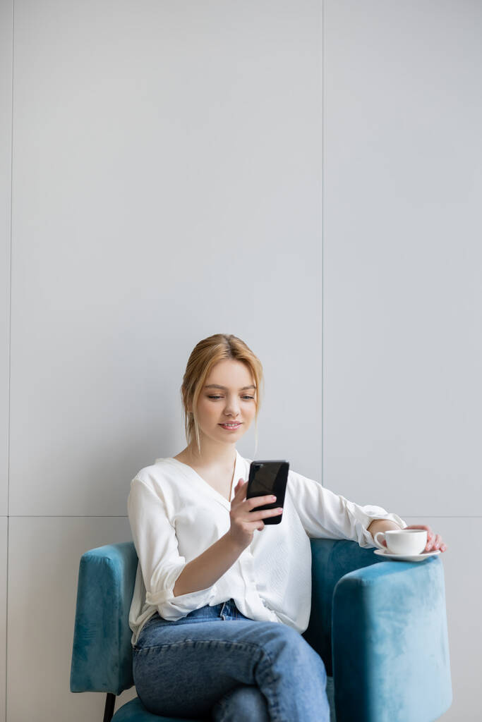 Giovane donna utilizzando smartphone e tenendo il caffè sulla poltrona a casa - Foto, immagini