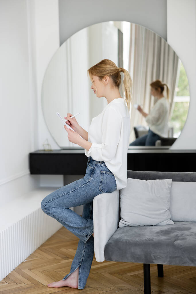 Side näkymä nuori blondi nainen kirjoittaa muistikirjaan lähellä sohvalla kotona  - Valokuva, kuva