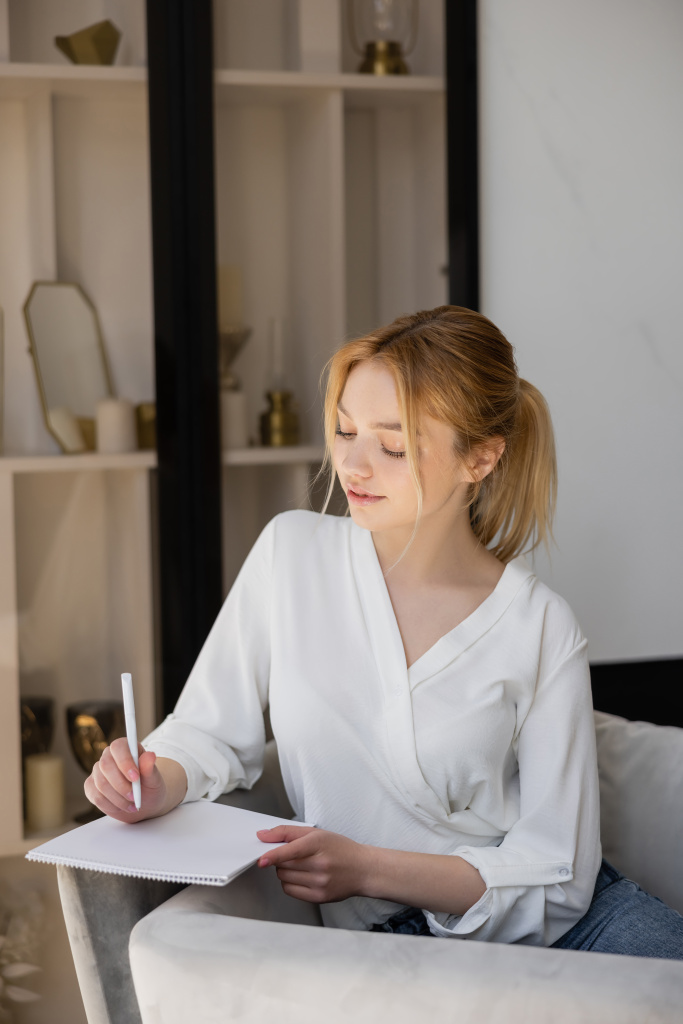 Csinos fiatal nő ír jegyzetfüzetbe, miközben otthon ül a karosszéken.  - Fotó, kép