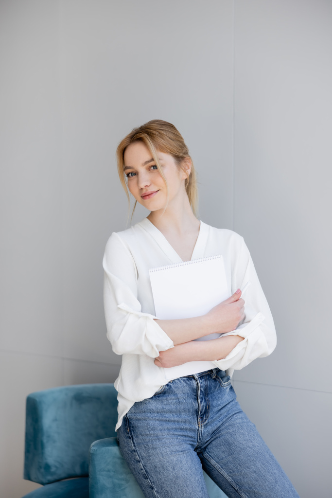 Blonde woman in blouse and jeans holding notebook near armchair at home  - Photo, Image