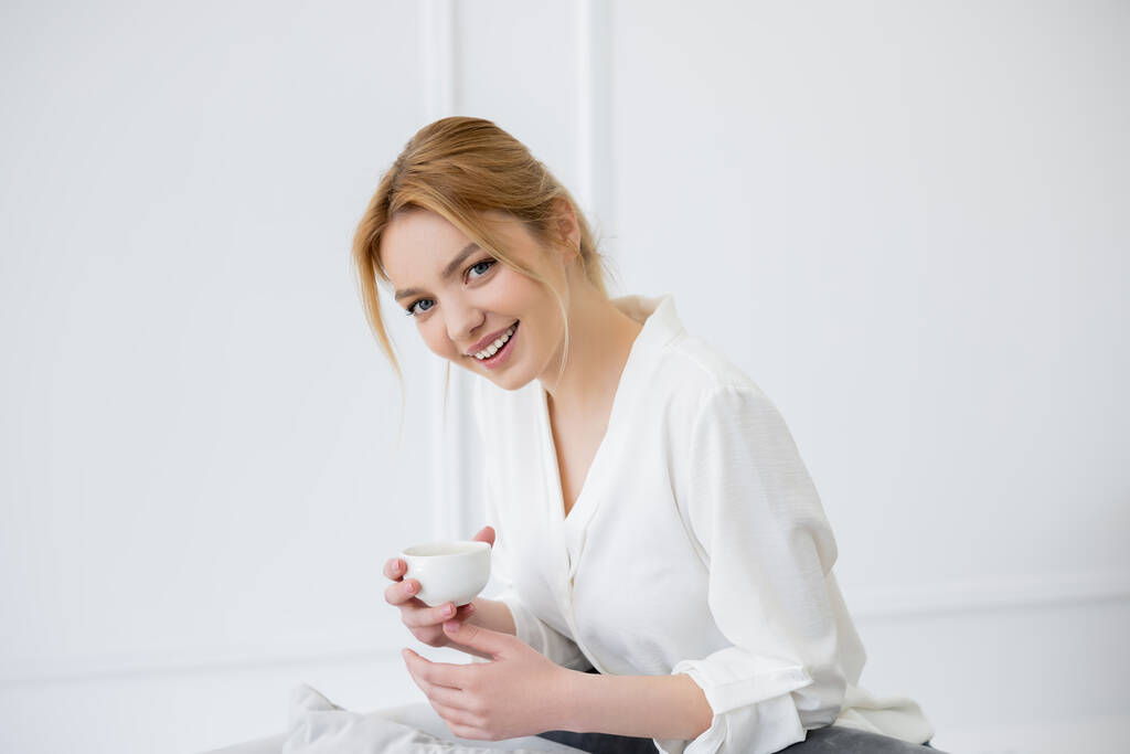 Счастливая молодая женщина в блузке держит чашку кофе и смотрит в камеру дома  - Фото, изображение
