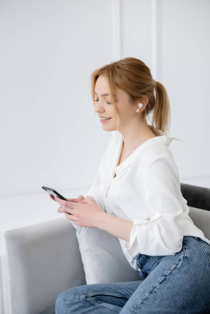 Mujer joven positiva en auriculares usando teléfono inteligente en la sala de estar  - Foto, imagen