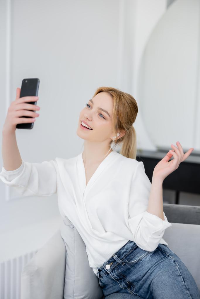 Mujer joven positiva en blusa y auricular que tiene videollamada en teléfono inteligente en la sala de estar  - Foto, imagen