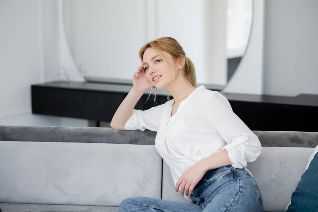 Vrolijke vrouw in witte blouse zittend op de bank in de woonkamer  - Foto, afbeelding