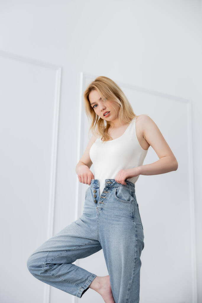 Jonge vrouw in bodysuit aanraken jeans en kijken naar de camera in de woonkamer  - Foto, afbeelding