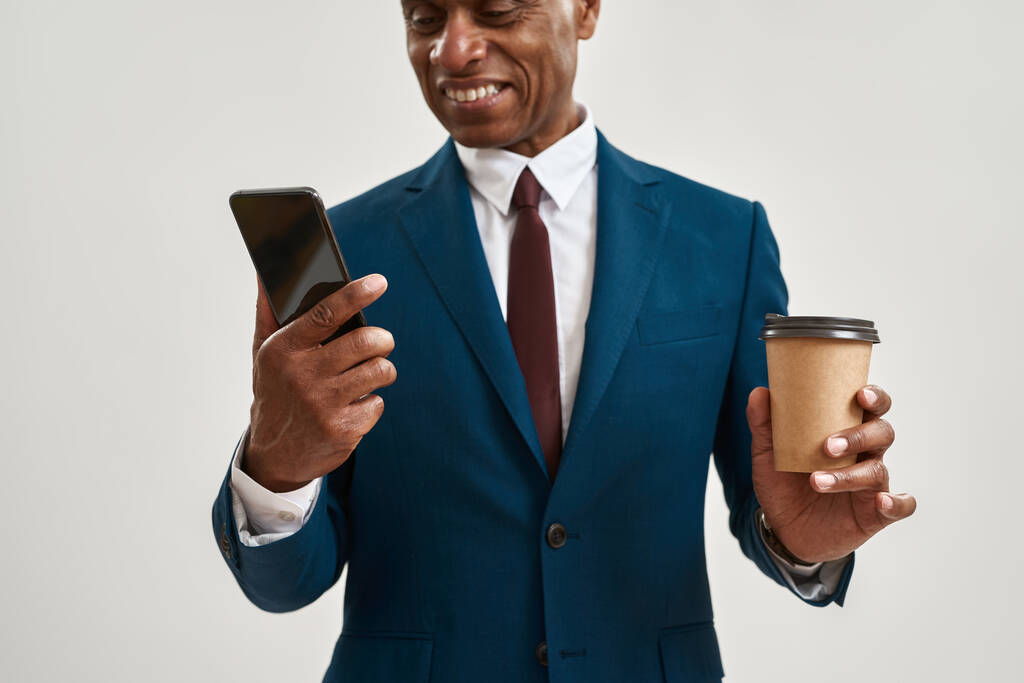 Kahveli, akıllı telefonlu, gülümseyen siyah CEO yöneticisi. Resmi kıyafet giyen kel bir yetişkin. Modern başarılı erkek yaşam tarzı kavramı. Beyaz arka planda izole edilmiş. Stüdyo çekimi. Boşluğu kopyala - Fotoğraf, Görsel