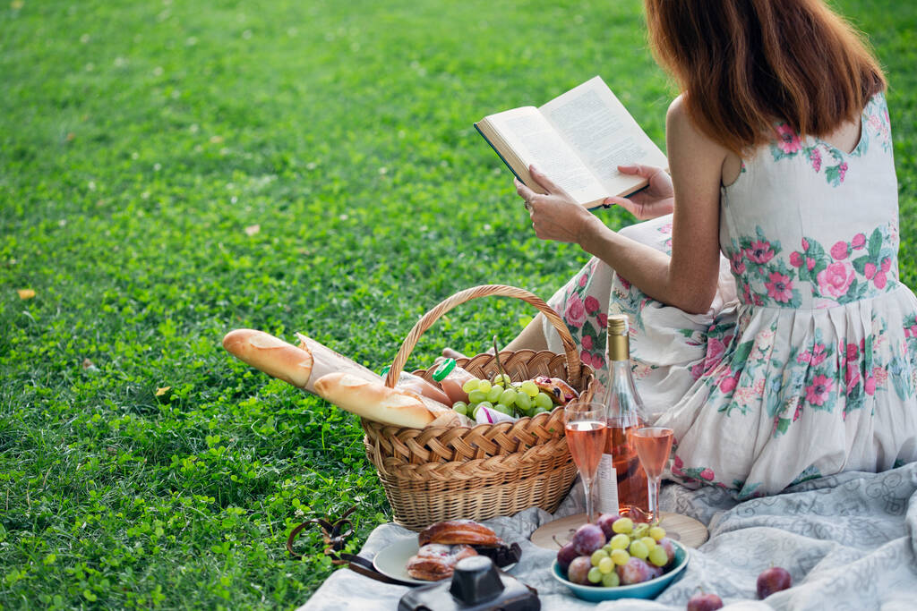 Summer - picnic in the meadow.  girl sitting reading a book and near a picnic basket and baguette, wine, glasses, grapes and roll - Photo, Image