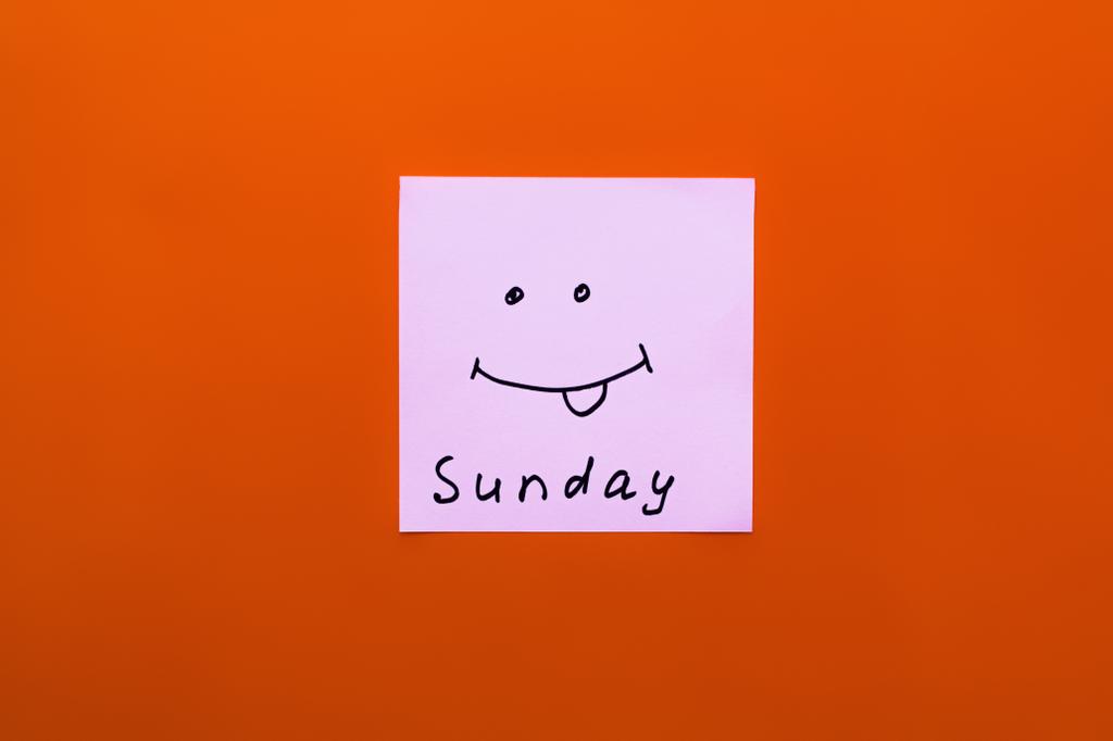日曜日のレタリングのある紙とオレンジの背景に舌を突き出す陽気な笑顔のトップビュー - 写真・画像