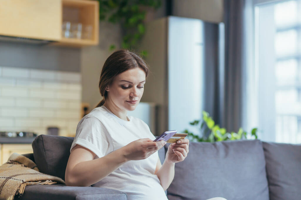 Maternity leave. Pregnant beautiful woman sitting on the couch at home, holding a mobile phone and credit card, in home clothes, ordering baby products online, choosing, buying clothes and toys for the baby. - Photo, Image