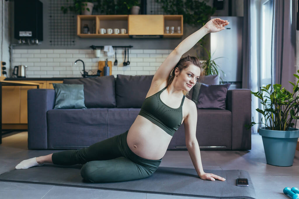 Embarazada hermosa mujer entra para los deportes en casa, se sienta y se encuentra en una alfombra deportiva en un uniforme deportivo, hace ejercicios, está satisfecho - Foto, imagen