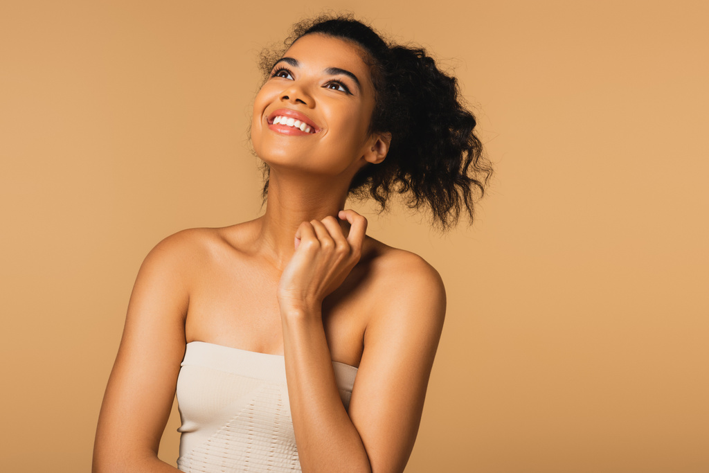 heureuse jeune femme afro-américaine aux épaules nues regardant isolée sur beige  - Photo, image