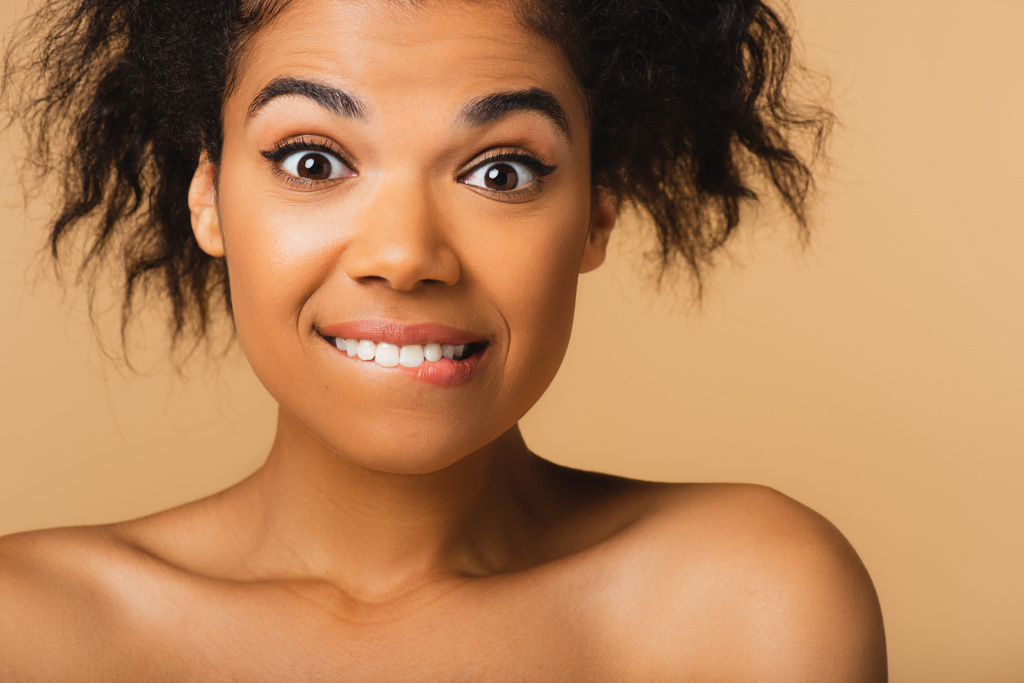mujer americana africana rizada con los hombros desnudos mordiendo labio aislado en beige  - Foto, Imagen