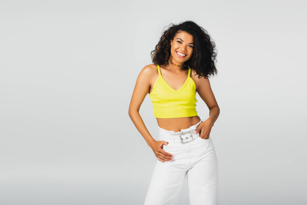 happy african american woman in yellow crop top and white pants posing with hand in pocket isolated on grey  - Photo, Image