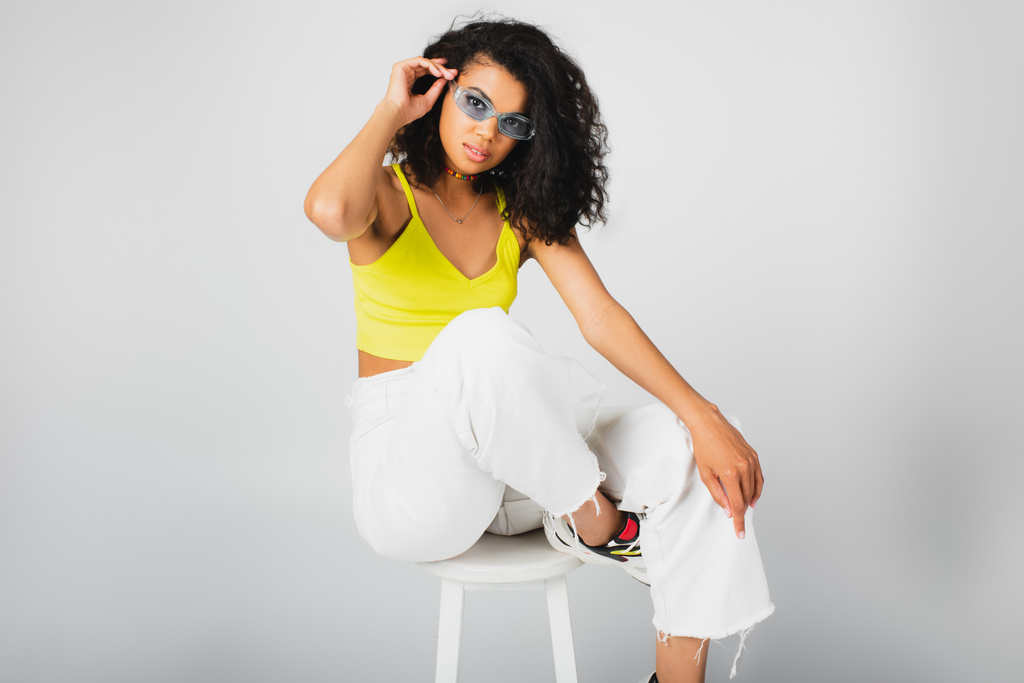 кучерява афроамериканська жінка в стильних сонцезахисних окулярах і модному вбранні позує на білому високому стільці на сірому
 - Фото, зображення