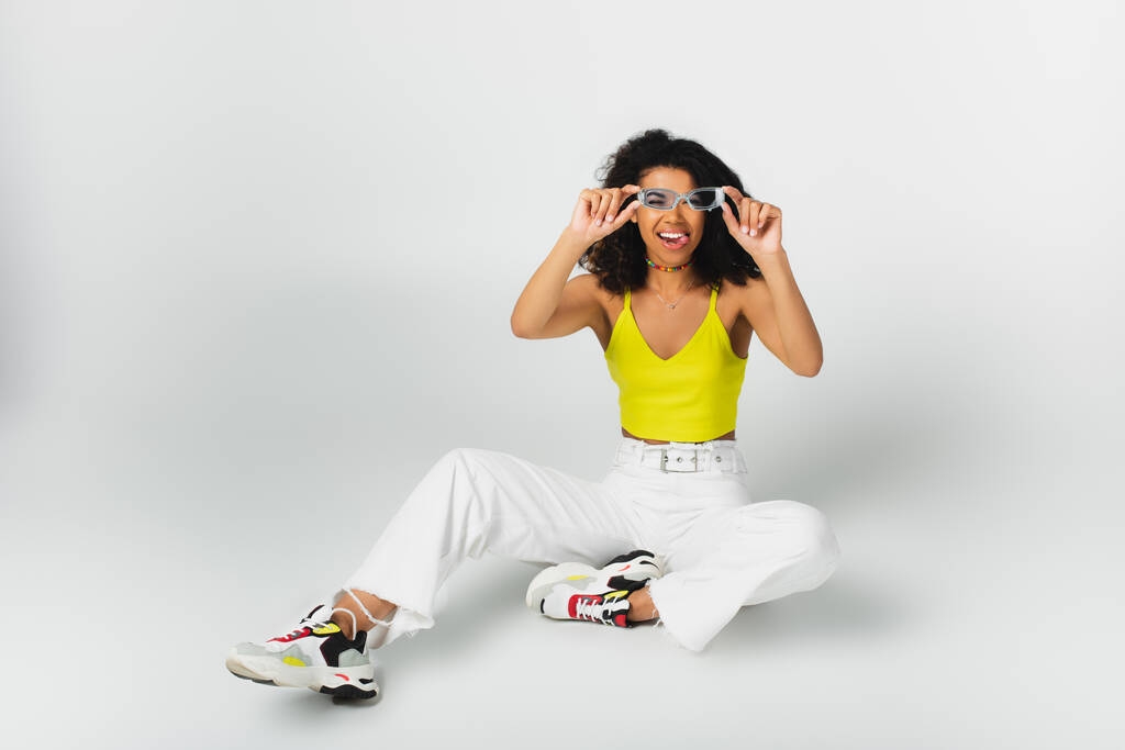 joyful african american woman adjusting blue sunglasses and sticking out tongue while sitting on grey - Photo, Image