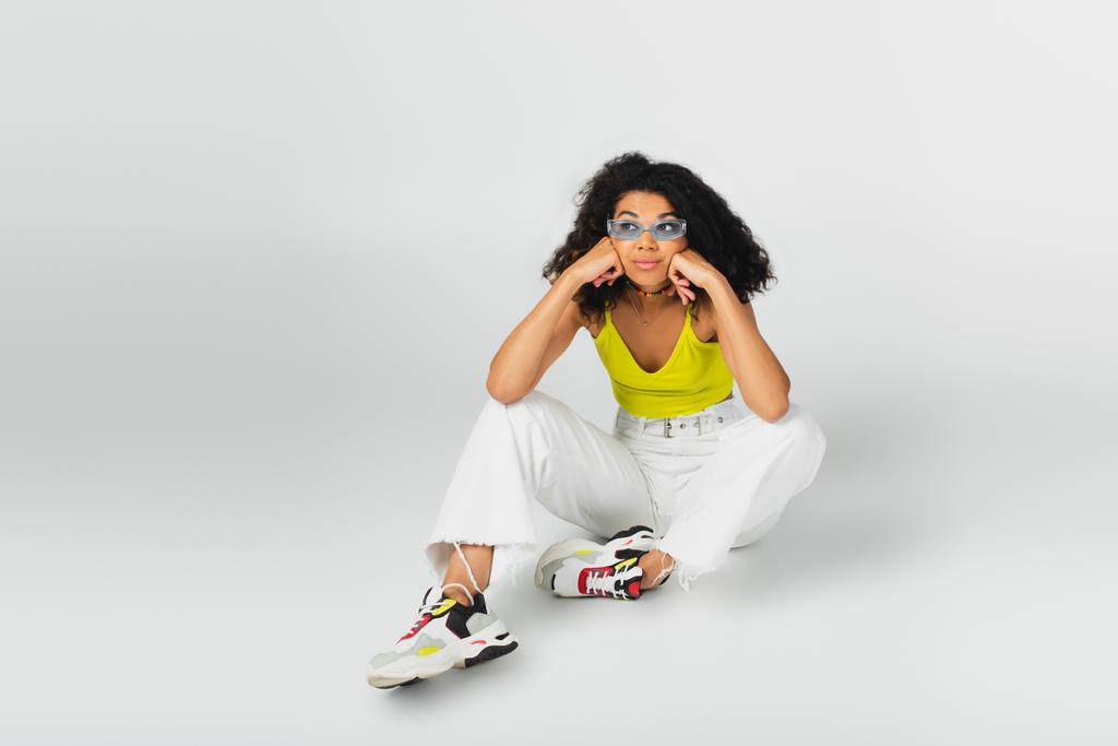 σγουρά αφροαμερικάνικη γυναίκα με μπλε γυαλιά ηλίου και μοντέρνα sneakers σε γκρι - Φωτογραφία, εικόνα