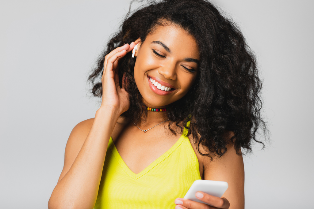 szczęśliwy Afryki amerykanka kobieta w bezprzewodowy słuchawki gospodarstwa smartfon odizolowany na szary  - Zdjęcie, obraz