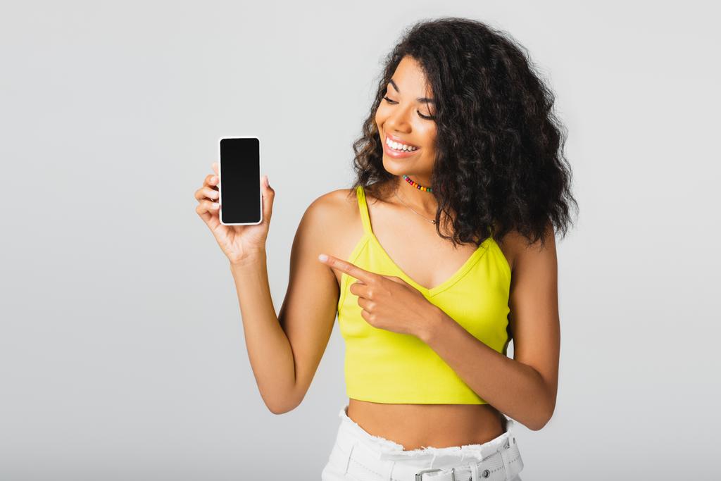 Χαρούμενη Αφροαμερικανή γυναίκα που δείχνει το smartphone με λευκή οθόνη απομονωμένη σε γκρι  - Φωτογραφία, εικόνα