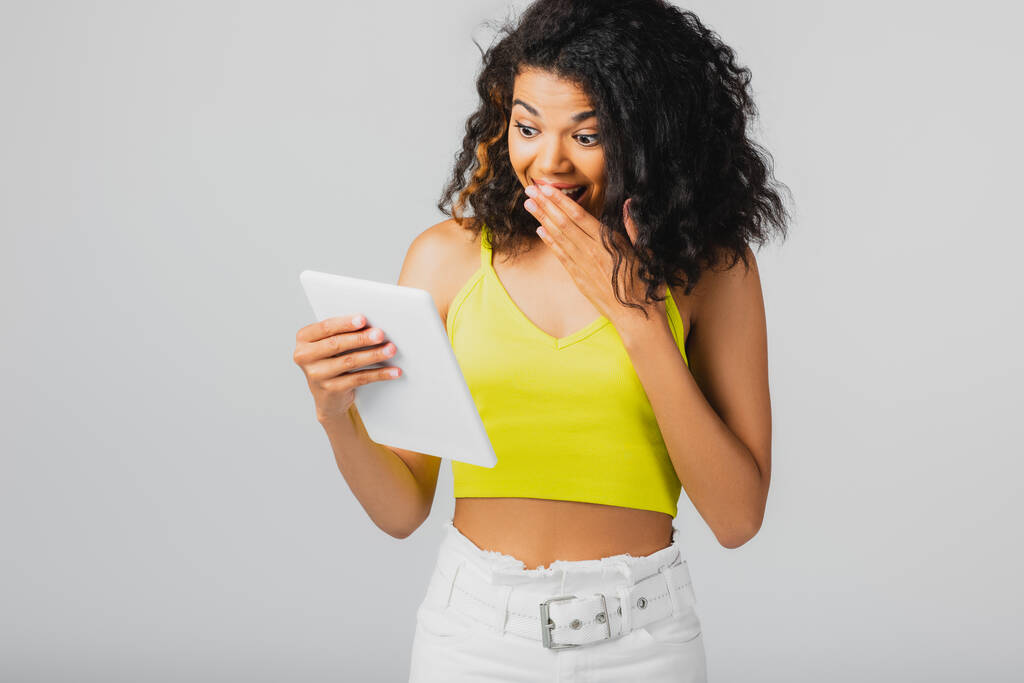 yllättynyt afrikkalainen amerikkalainen nainen keltainen sato alkuun katsomalla digitaalinen tabletti eristetty harmaa - Valokuva, kuva