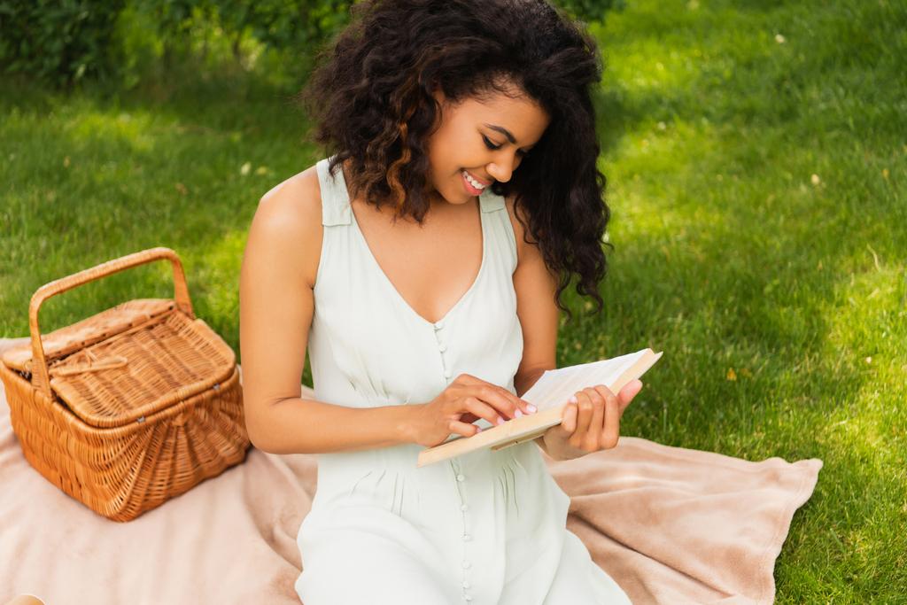 χαρούμενη Αφροαμερικανή γυναίκα σε βιβλίο ανάγνωσης φορεμάτων και κάθεται σε κουβέρτα κοντά στο καλάθι πικνίκ στο πάρκο - Φωτογραφία, εικόνα
