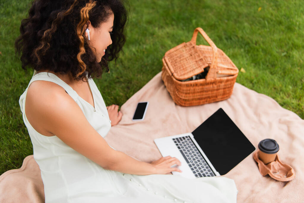 кудрявая африканская американка с ноутбуком возле плетеной корзины и смартфоном на одеяле в парке  - Фото, изображение