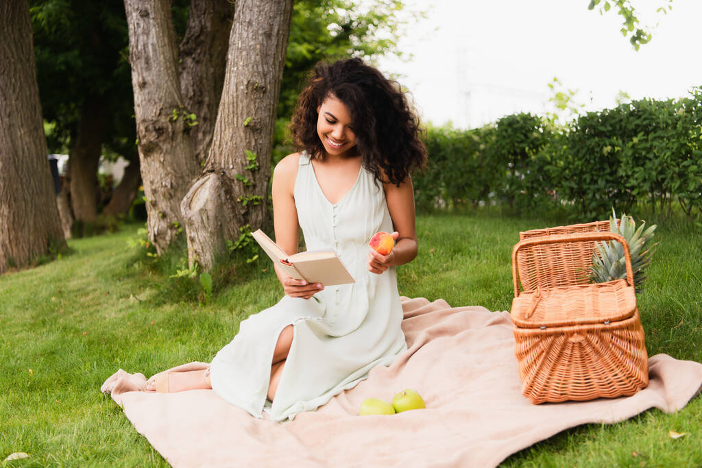 heureuse femme afro-américaine en robe blanche tenant pêche et livre de lecture pendant le pique-nique  - Photo, image