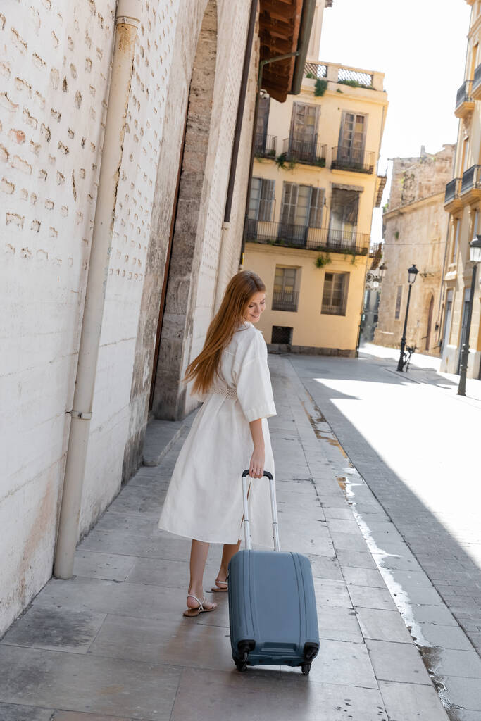 Valencia 'da sokakta bavulla yürüyen, gülümseyen genç bir kadın.  - Fotoğraf, Görsel