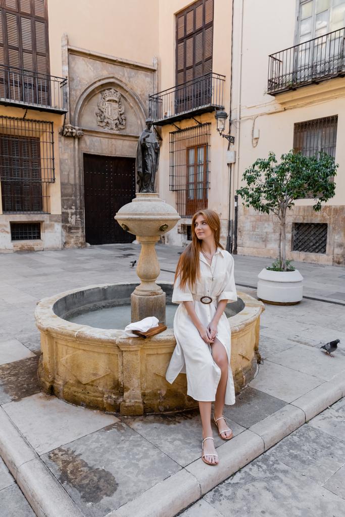 pełna długość ruda kobieta w białej sukience siedzi w pobliżu fontanny na ulicy Walencji - Zdjęcie, obraz