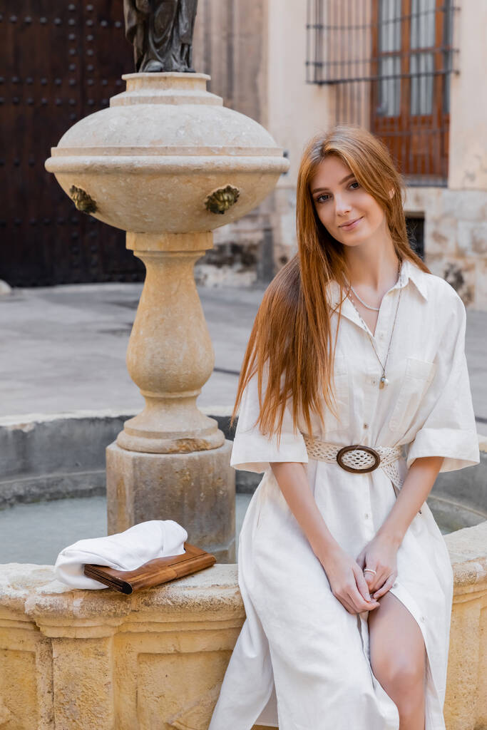 pelirroja sonriente en vestido blanco sentada cerca de la fuente en la calle de valencia - Foto, Imagen