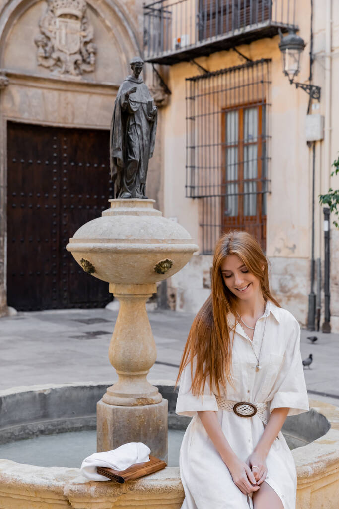 vrolijk en roodharige vrouw in witte jurk zitten in de buurt fontein op straat van valencia - Foto, afbeelding