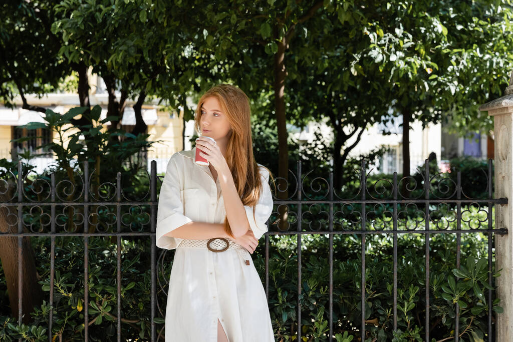 young redhead woman in dress holding coffee to go near fence in european green park - Photo, Image