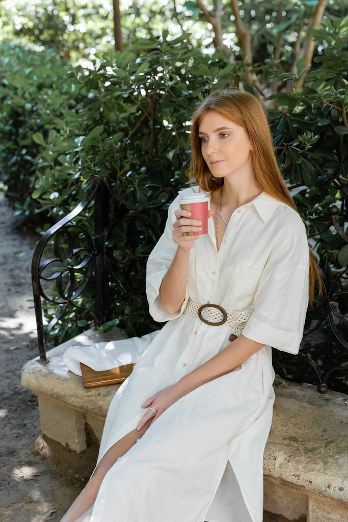 jeune rousse femme en robe tenant tasse en papier et assis sur un banc en béton dans le parc vert - Photo, image