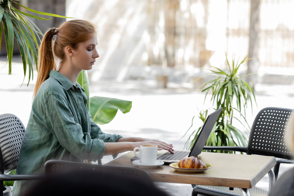 vista laterale di freelance con capelli rossi utilizzando computer portatile vicino a tazza di caffè e gustoso croissant in terrazza caffè - Foto, immagini