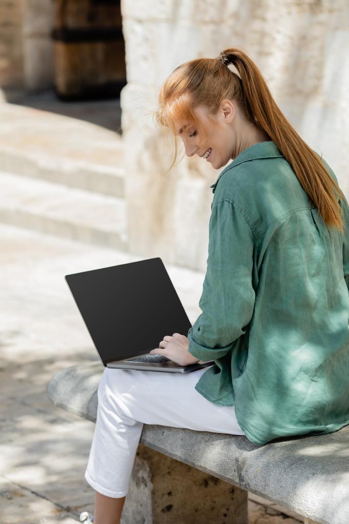uśmiechnięty freelancer siedzi na betonowej ławce i za pomocą laptopa z pustym ekranem - Zdjęcie, obraz