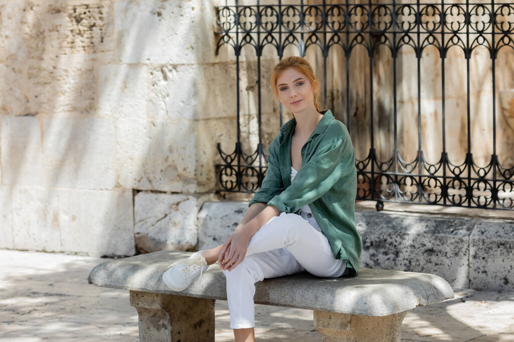 young woman with red hair in green linen shirt sitting on concrete bench near forged fence - Photo, Image