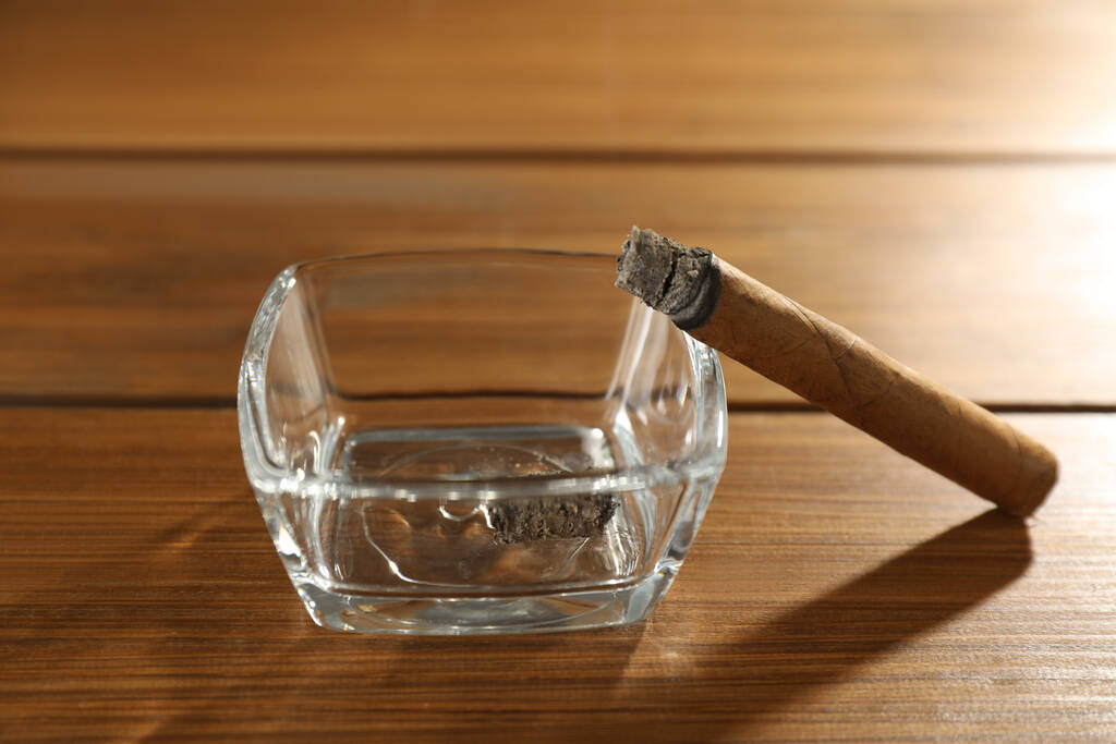 Smoldering πούρο κοντά σε γυάλινο τασάκι σε ξύλινο τραπέζι, closeup - Φωτογραφία, εικόνα