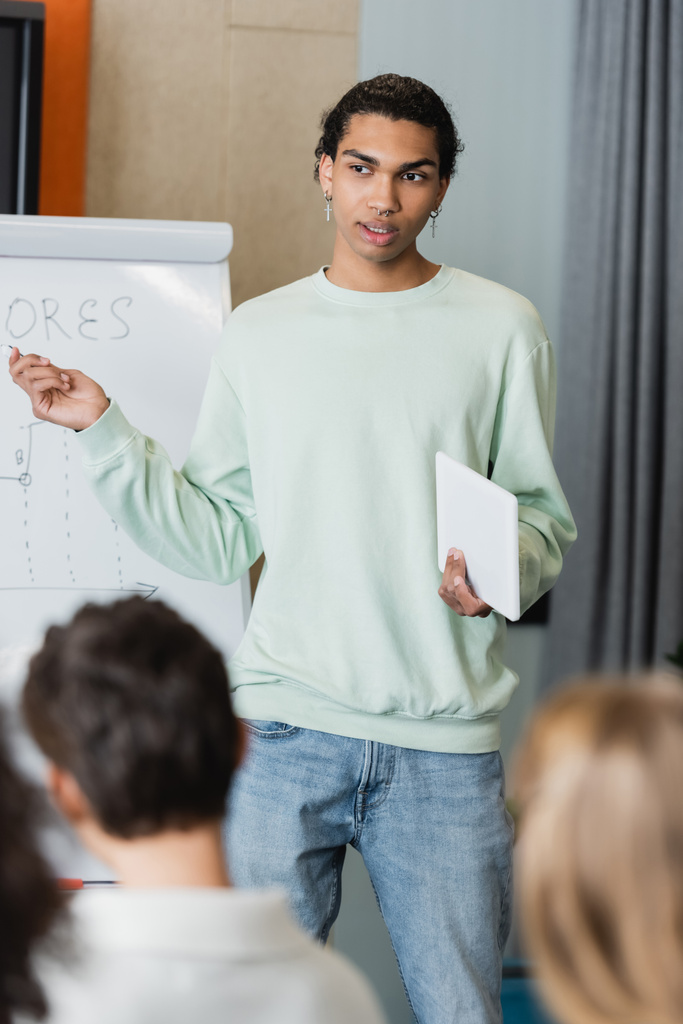Afrykański amerykański student z tabletem cyfrowym rozmawia z niewyraźnymi kolegami z klasy w pobliżu tablicy z zadaniem  - Zdjęcie, obraz
