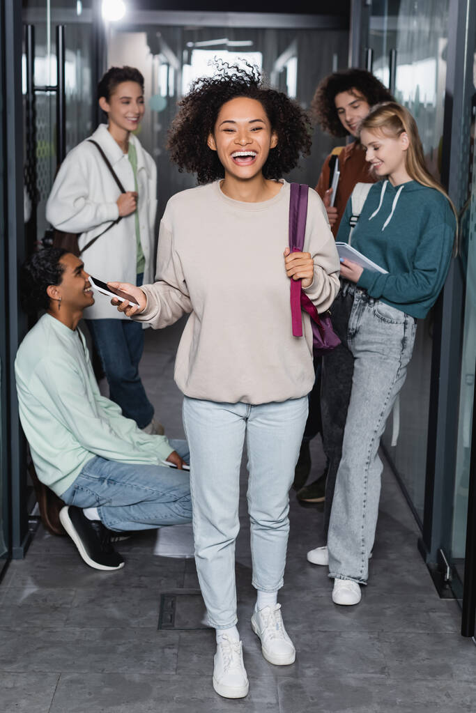 comprimento total de mulher afro-americana rindo com smartphone perto de amigos inter-raciais no corredor da universidade - Foto, Imagem