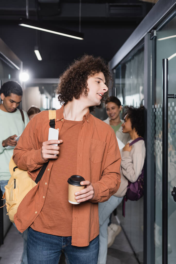 kudrnatý student s nápojem a batohem odvracející se v blízkosti multikulturní spolužáci na rozmazaném pozadí - Fotografie, Obrázek