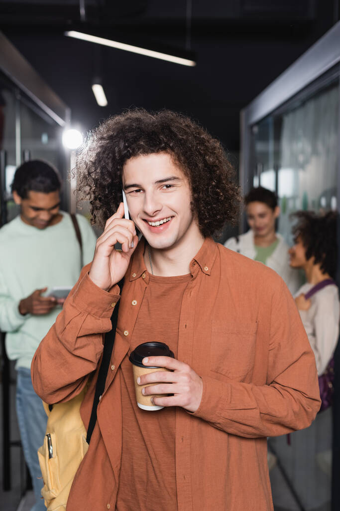 étudiant bouclé et heureux avec tasse en papier parlant sur smartphone près de personnes multiethniques floues - Photo, image