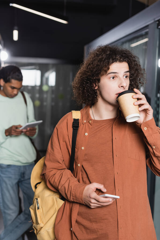 uomo riccio con cellulare bere caffè vicino studente americano africano utilizzando tablet digitale su sfondo sfocato - Foto, immagini