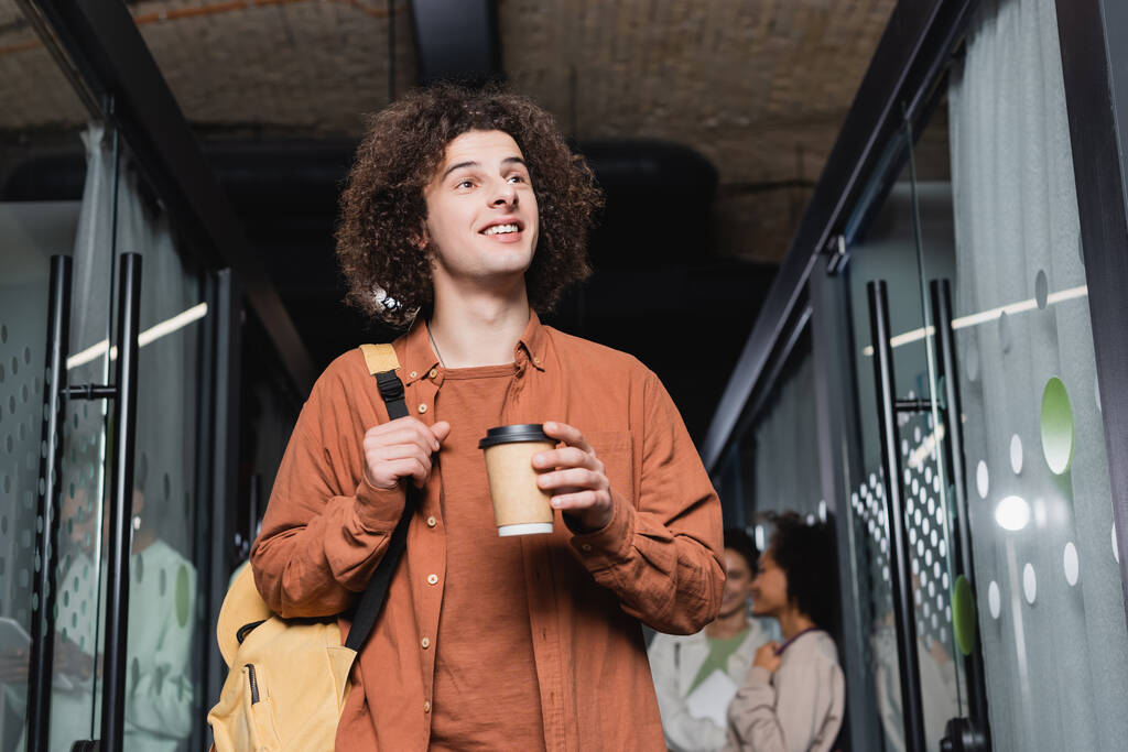 kudrnatý student s kávou jít a batoh dívá pryč a usmívá se na univerzitě - Fotografie, Obrázek