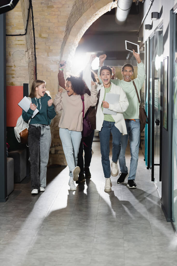 pełna długość podekscytowanych wielonarodowych studentów spacerujących korytarzem i pokazujących gest wygranej - Zdjęcie, obraz