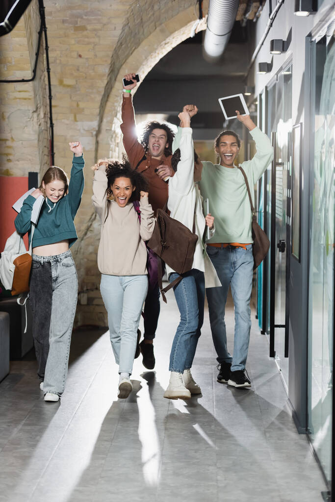 веселые мультиэтнические студенты с рюкзаками и гаджетами демонстрируют победный жест в университетском коридоре - Фото, изображение