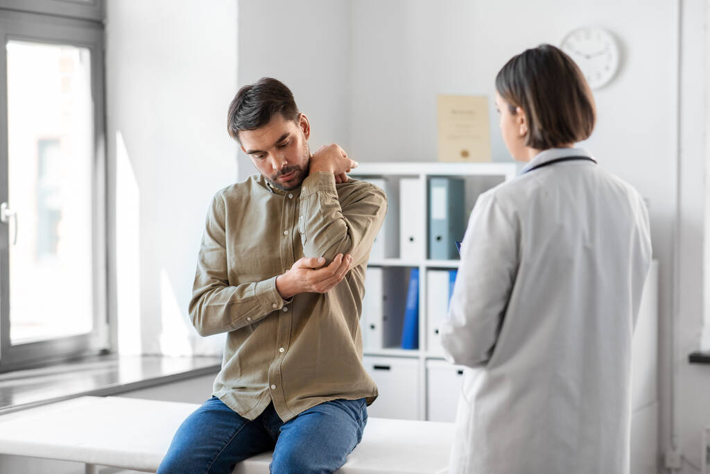 lääketiede, terveydenhuolto ja ihmisten käsite - miespotilas osoittaa kipeää käsivartta naislääkärille sairaalan lääketieteellisessä toimistossa - Valokuva, kuva