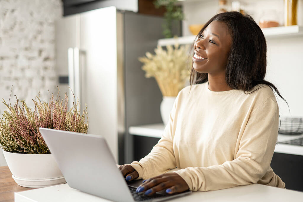 Inspiré afro-américaine pigiste femme appréciant le travail à distance de la maison, dame gaie en utilisant ordinateur portable assis dans la cuisine à la maison, regarde loin rêveur - Photo, image