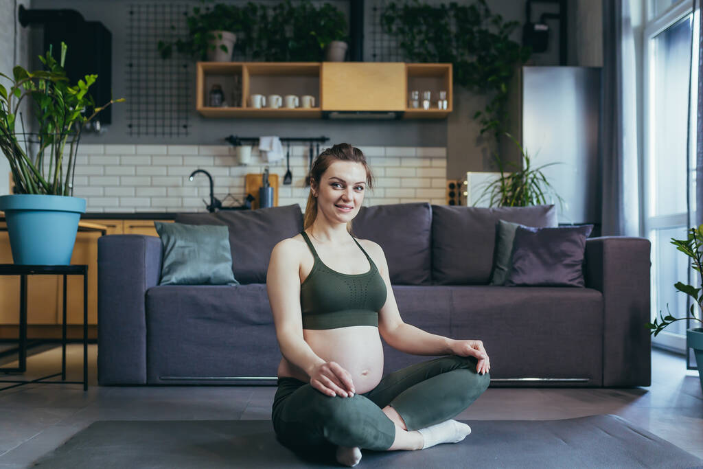 Una mujer embarazada medita y practica yoga en una posición de loto en casa - Foto, imagen