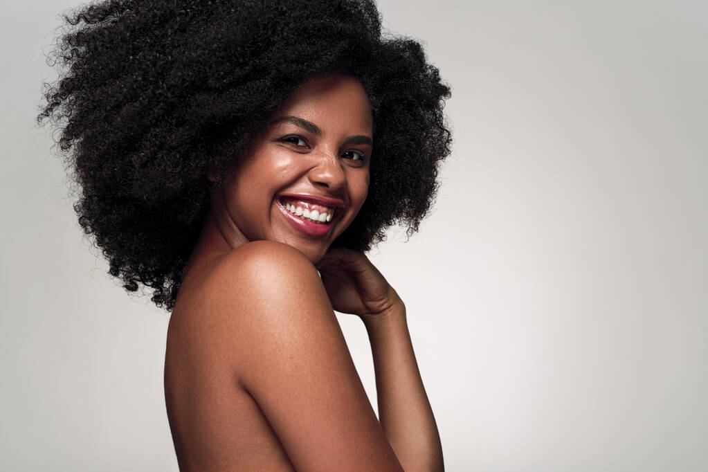 Side view of cheerful African American woman with bare shoulders and curly hair looking at camera, with smile and touching face during skin care routine against gray background - Photo, Image