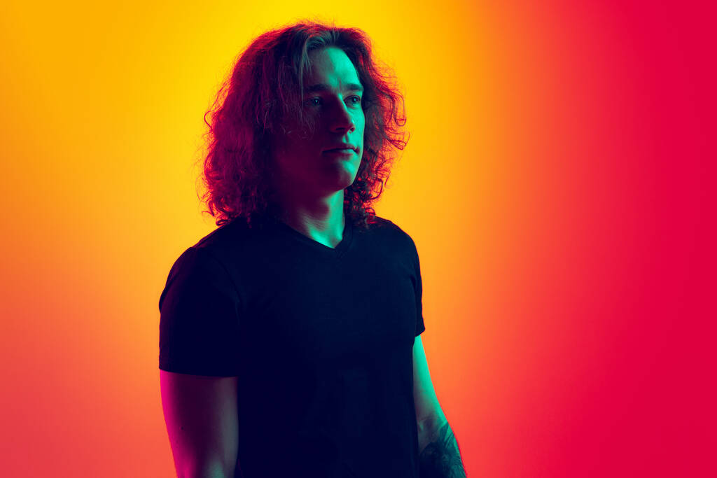 Thinking. Young serious man with long curly hair posing isolated on gradient yellow-magenta background in neon. Concept of beauty, fashion, youth culture and emotions. Half-length male portrait. - Photo, Image