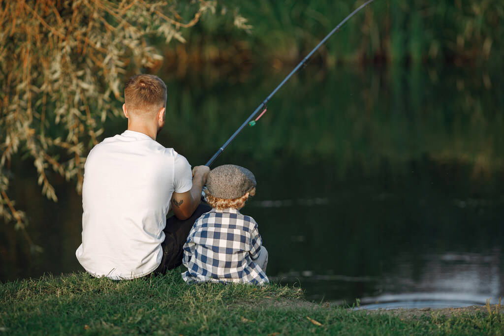 Il giovane papà e il suo bambino passano del tempo all'aperto insieme. Un bambino riccio che indossa una camicia a quadri e un cappello. Ragazzo seduto vicino al fiume e pesca con suo padre. - Foto, immagini
